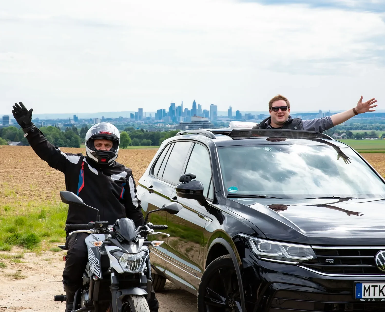 Zwi Fahrlehrer winken aus dem Auto und vom Motorrad. Frankfurter Skyline im Hintergrund