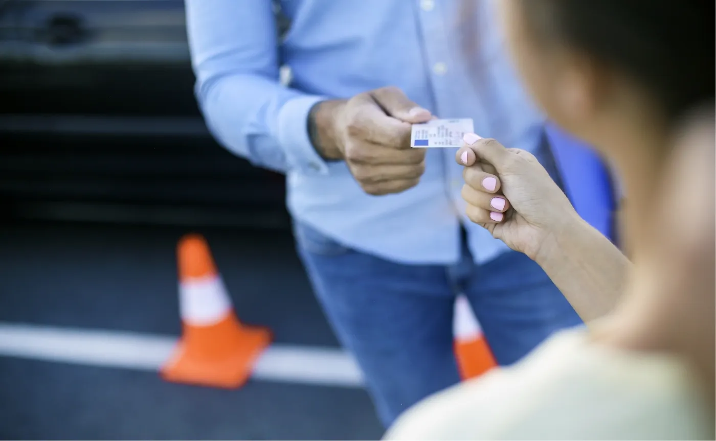 ein Fahrlehrer übergibt einer Fahrschülerin mit rosa lackierten Fingernägeln ihren Führerschein